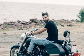 Fototapeta na wymiar handsome bearded motorcyclist on black motorcycle at sandy beach near sea