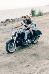 Plakat young couple of bikers riding black motorcycle at sandy beach