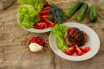a grilled chicken thigh lies with a lettuce on a plate. On a plate are cut tomatoes, dill and lettuce. whole cucumbers lie on a canvas napkin. dinner in nature.