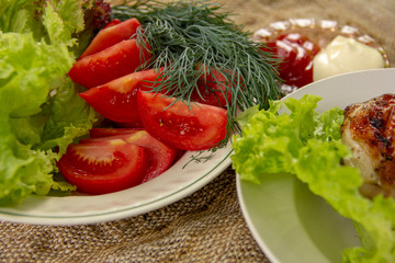 a grilled chicken thigh lies with a lettuce on a plate. On a plate are cut tomatoes, dill and lettuce. whole cucumbers lie on a canvas napkin. dinner in nature.
