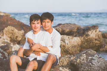 Fototapeta premium Homosexual portrait young asian couple sitting hug together on rock or stone in the beach in summer, asia gay going tourism for leisure and relax with happy in vacation at sea, LGBT legal concept.