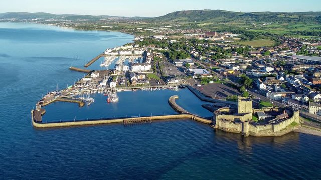 Medieval Norman Castle in Carrickfergus near Belfast in sunrise light. Aerial 4K revealing video with marina, yachts, parking, town and far view of Belfast in the background