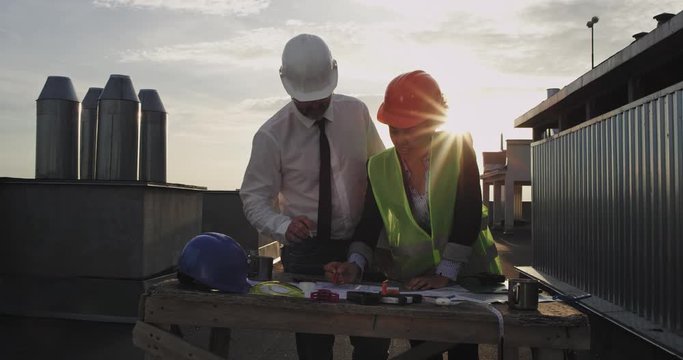 The African American young lady is wearing a orange colored construction helmet and a bright yellow jacket the man is wearing a white shirt with a white construction helmet as they both work on some