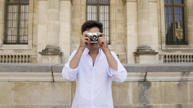 PARIS, FRANCE, APRIL 2019. Gimbal slow motion middle shot of happy young man in white shirt and eyeglasses making a photo with a film camera on background of Louvre museum