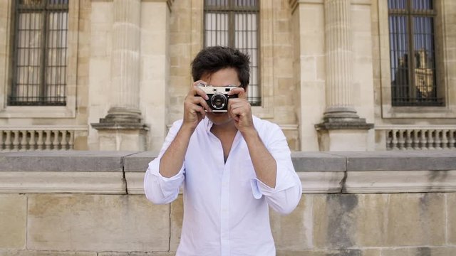 PARIS, FRANCE, APRIL 2019. Gimbal middle shot of happy young man in white shirt and eyeglasses with a film camera making a photo straight at camera on background of Louvre museum