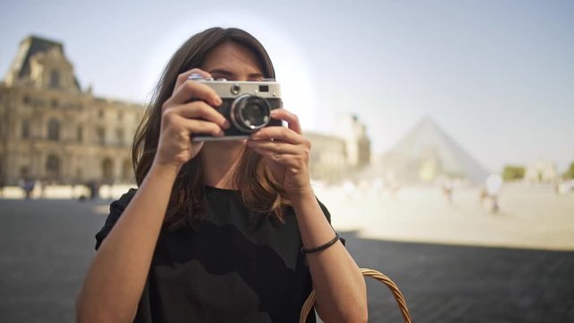 PARIS, FRANCE, APRIL 2019. Gimbal slow motion portrait shot of young caucasian brunette woman making photo with a film camera on background of blurred Louvre museum and glass pyramid