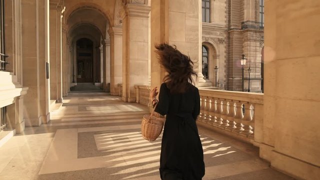 PARIS, FRANCE, APRIL 2019. Slow motion of back of woman in black dress running alone holding a wicker basket outside Louvre museum sunshine rays