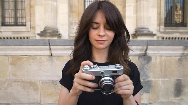 Gimbal slow motion middle shot of young woman looking at the camera rewinding a film camera making photo on background of old European city