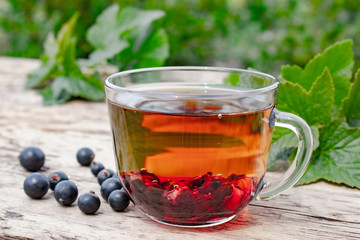 Glass cup of fruit tea with blackcurrant berries on a wooden table near green leaves and berries of currant.