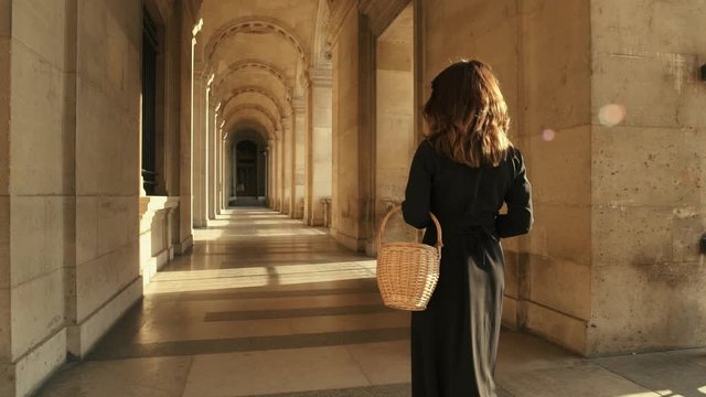 PARIS, FRANCE, APRIL 2019. Slow motion of back of woman in black dress walking alone holding a wicker basket outside Louvre museum sunshine rays