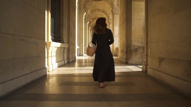 PARIS, FRANCE, APRIL 2019. Slow motion of back of woman in black dress walking alone holding a wicker basket outside Louvre museum sunshine rays