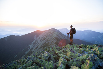 caucasian man hiker with backpack and trekking sticks is on the peak of green stones mountain enjoy a panoramic view of mountains in the sunset light