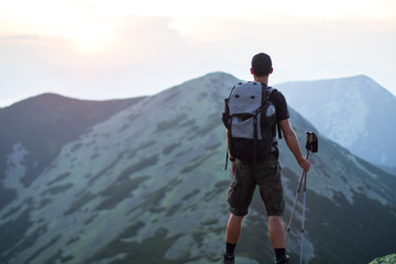 caucasian man hiker with backpack and trekking sticks is on the peak of green stones mountain enjoy a panoramic view of mountains in the sunset light