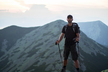 caucasian man hiker with backpack and trekking sticks is on the peak of green stones mountain enjoy a panoramic view of mountains in the sunset light