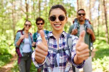 travel, tourism, hike and people concept - group of friends with backpacks and asian woman showing thumbs up gesture in forest