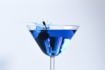 Cocktail with blue liquid in glass. Glass with blue water pouring with liquid with splashes and drops. Martini glass filling with alcohol with splashes on white background. Refreshing drink concept.