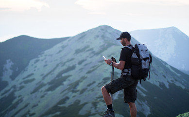 caucasian man hiker with backpack and trekking sticks is on the peak of green stones mountain is searching something in the smartphone