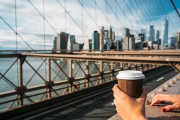 Tuinposter Detail of the hand of a girl holding a glass of coffee while watching New York from a bridge © ManuPadilla