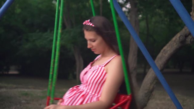 Pregnant girl in the park swinging on a swing. Happy girl with long dark hair in a striped white-red dress on a swing n the park.