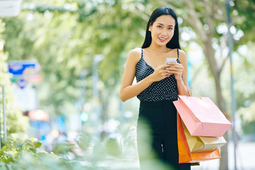 Portrait of Asian brunette in beautiful suit with shopping bags using mobile phone while walking along the street and smiling at camera