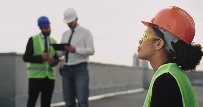 On construction site thinking young African woman engineer on the rooftop of building wearing a orange helmet and yellow glasses closeup to the camera , background two workers analyzing something