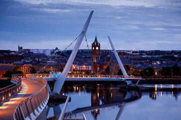 Derry, Ireland. Illuminated Peace bridge in Derry Londonderry, City of Culture, in Northern Ireland with city center at the background. Night cloudy sky with reflection in the river at the dusk - obrazy, fototapety, plakaty