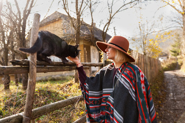Black cat and stylish traveling woman wearing  authentic boho chic style poncho and hat near old wooden countryside buildings Woman have a fun in autumn time.Travel and wanderlust concept.