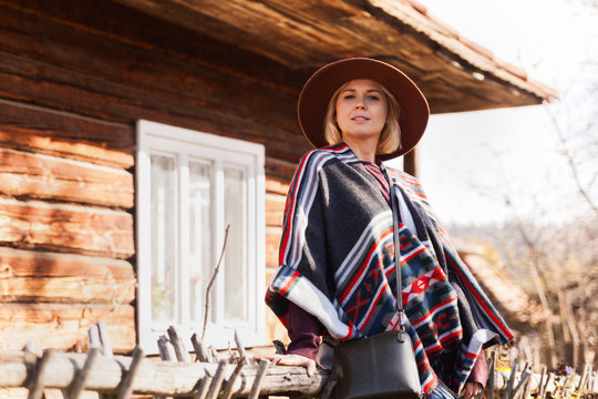 Stylish hipster traveling woman wearing  authentic boho chic style poncho and hat near old wooden countryside buildings Woman exploring nature in autumn time.Travel and wanderlust concept.