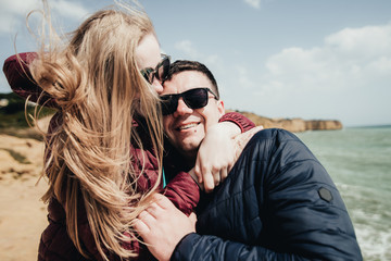 Happy young couple embracing and holding hands by the ocean view. Man and woman sitting together on the cliff. Smiling pair in black eyeglasses enjoying vacation in Portugal