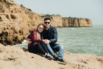 Happy young couple embracing and holding hands by the ocean view. Man and woman sitting together on the cliff. Smiling pair in black eyeglasses enjoying vacation in Portugal