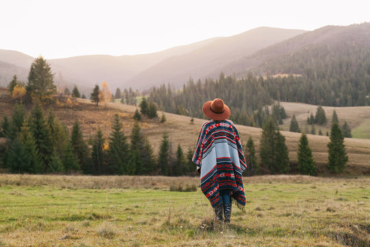 Stylish hipster traveling woman wearing  authentic boho chic style poncho and hat in autumn landscape. Happy young woman exploring sunny mountains. Travel and wanderlust concept.