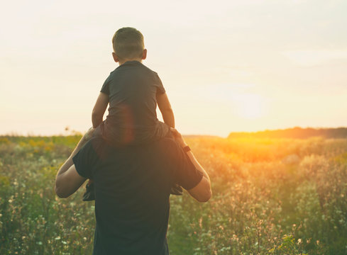 The son is sitting on the father's shoulders and they are looking on the sunset in the field