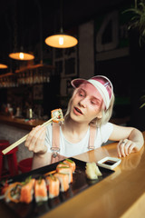Pretty girl in a pink cap and cute clothes holds a roll of sushi on sticks and is about to eat. Girl enjoys eating sushi in an Asian restaurant with an appetite looking at the roll in her hand.