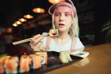 Surprised lady in pink cap holding chopsticks with sushi roll, looking surprised at food. Young girl holding a beautiful piece of sushi on chopsticks, close up photo. Background