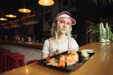 Portrait of a beautiful girl sitting at a table in a restaurant with a plate of sushi rolls, holding chopsticks in her hands, looking into the camera with a serious face. Cute lady dines for sushi