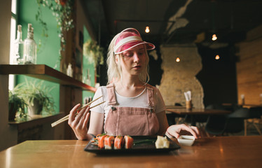 Attractive girl sits in a cozy Japanese restaurant with a plate of sushi rolls and soy sauce, holds chopsticks, looks downstairs, wears a pink cap and sundress. Asian restaurant concept.