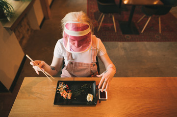 Attractive girl sits in a cozy Japanese restaurant with a plate of sushi rolls and soy sauce, holds chopsticks, looks downstairs, wears a pink cap and sundress. Asian restaurant concept. Top view