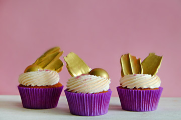 Sweet organic cupcake in purple wrap with cream cheese frosting swirl and golden chocolate decoration on white wooden textured table. Close up, copy space, background.