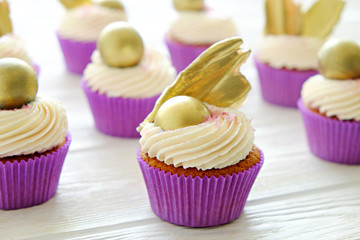 Sweet organic cupcake in purple wrap with cream cheese frosting swirl and golden chocolate decoration on white wooden textured table. Close up, copy space, background.