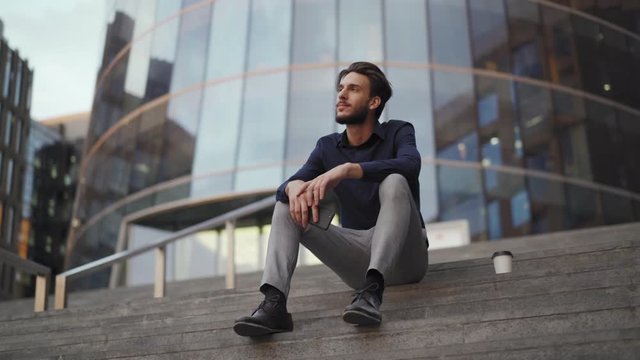 Panning shot of relaxed young businessman sitting on steps of modern office building with cell phone in his hands and looking away dreamily