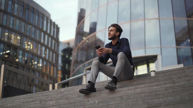 Panning shot of handsome young businessman sitting on steps of modern office building with smartphone in his hands and looking away thoughtfully