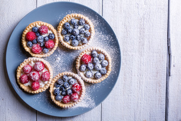 Cakes with raspberries and blueberries on a gray large plate sprinkled with icing sugar on a white wooden background Top view