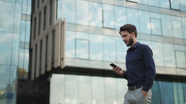 Tracking zoom in shot of handsome young businessman text messaging on cell phone while walking down city street in slow motion with hand in pocket. Man looking forward and away thoughtfully