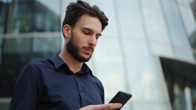 Zoom in slow motion shot of handsome young businessman text messaging on cell phone while standing in city street. Man looking away thoughtfully