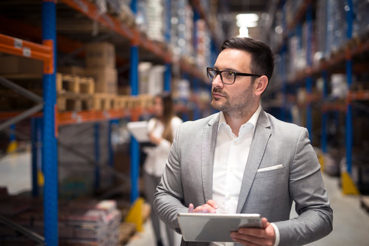 Manager visiting warehouse. Portrait of successful businessman manager CEO holding tablet and walking through warehouse storage area looking towards shelves.