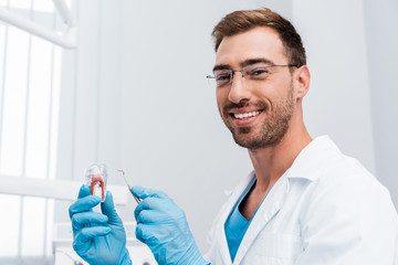 handsome bearded dentist holding dental instrument and tooth model while smiling in clinic