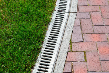 iron drainage system between paving slabs and green lawn, closeup of an iron grille along a pedestrian pavement. - obrazy, fototapety, plakaty