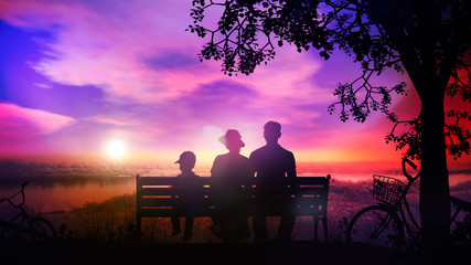 Fototapeta na wymiar Family rests on a bench under the tree and watches sunset over the river