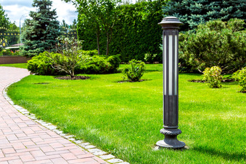 a garden ground lamp made of iron mounted on a green lawn in a park near a stone walkway for walks in a summer garden among a plant of bushes and trees.