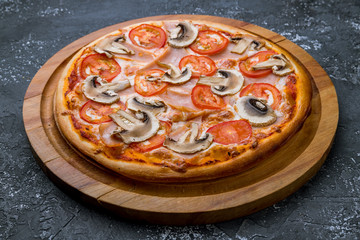 Pizza with ham and mushrooms and tomatoes on wooden board on black concrete